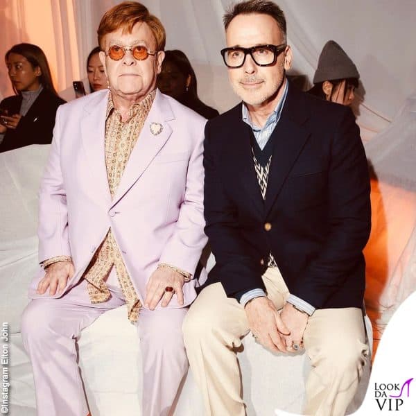 completo lilla, camicia fantasia e spilla a cuore per Elton John, al défilé con il marito David Furnish